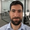 A man with a beard and short dark hair is standing in an office with glass walls and empty meeting rooms in the background.