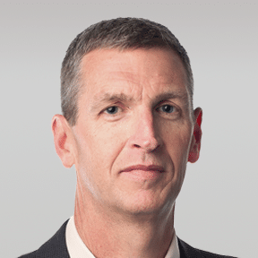 A middle-aged man with short brown hair wearing a dark suit, white shirt, and neutral expression, against a plain light background.